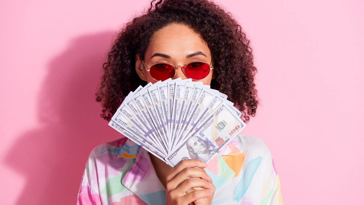 A woman folds a fan of $100 bills in front of her face in front of a pink background.