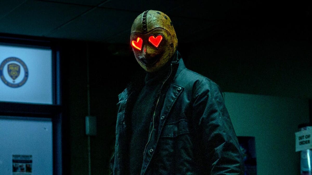 A person in all black, wears a mask with red hearts for eyes.