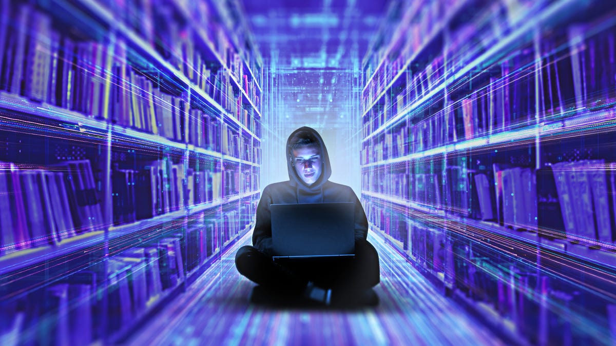 A man in a hoodie sits with a laptop, surrounded by rows of books