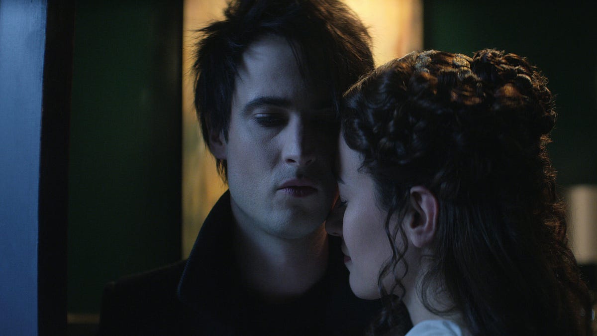 A man with great cheekbones and very black hair gazes down from a gothic throne.