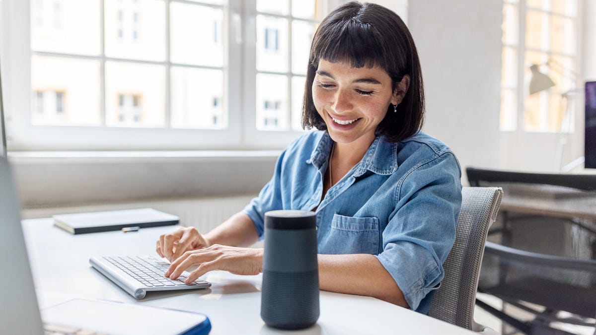 A woman at a computer desk smiles at a smart speaker beside her.