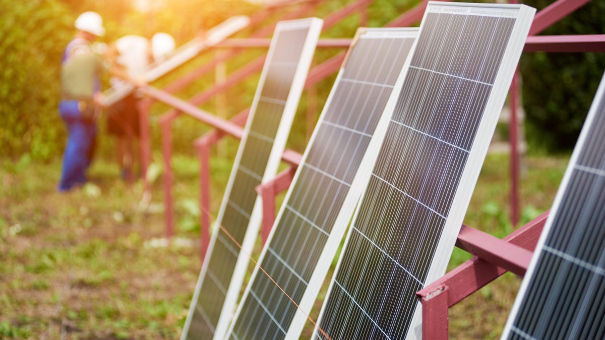 Solar panels leaning against a metal rack outdoors, person with a hard hat in the distance.