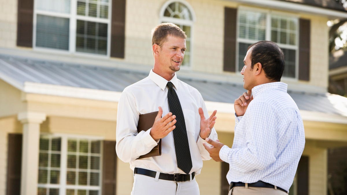 A salesperson in white talks to a homeowner outside a yellow home.