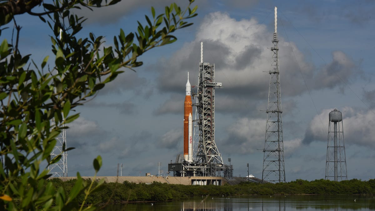 On the left, in the foreground, are leafy branches of a tree. At the bottom of the image, tracing all the way to the far-ground is a lake, in which the SLS rocket's reflection is seen. That's because the bright orange rocket itself, and the Orion spacecraft atop the rocket, is situated behind this lake. It's a cloudy day.