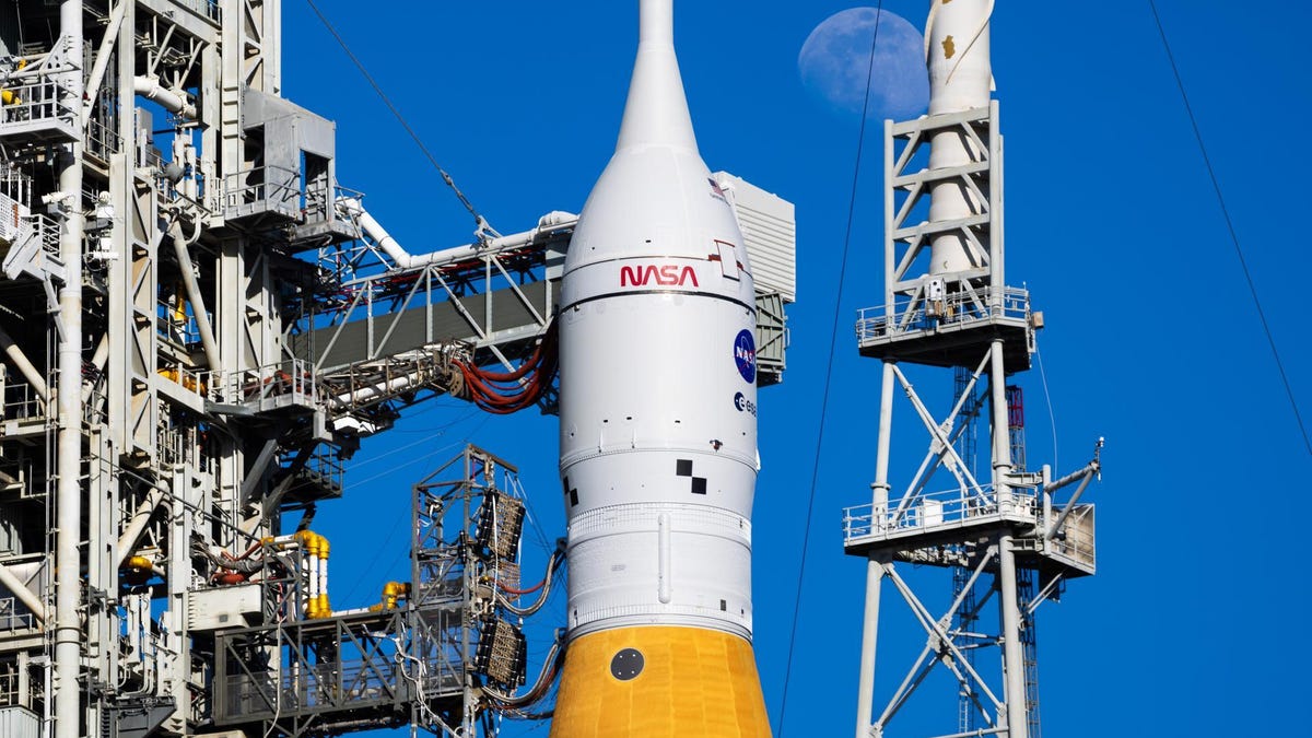 The Orion spacecraft atop the SLS rocket, with launch gantry alongside. The moon can be seen in the background in a blue sky.