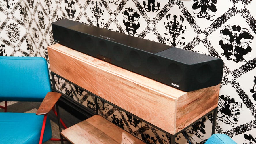 A black Sennheiser Ambeo Soundbar Max sits on a wooden console table in front of black and white patterned wallpaper.