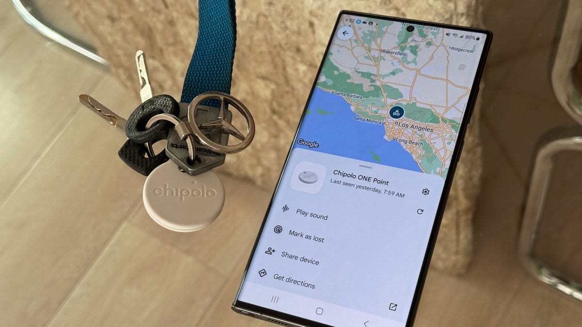 a Bluetooth tracker attached to a set of keys on a key ring sits on a tabletop next to a phone that shows its location