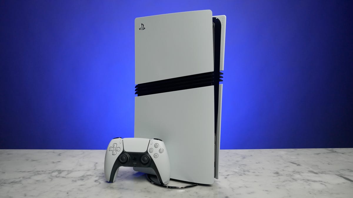 A PlayStation 5 Pro sits vertically on a table in front of a blue background. A gaming controller sits next to it.