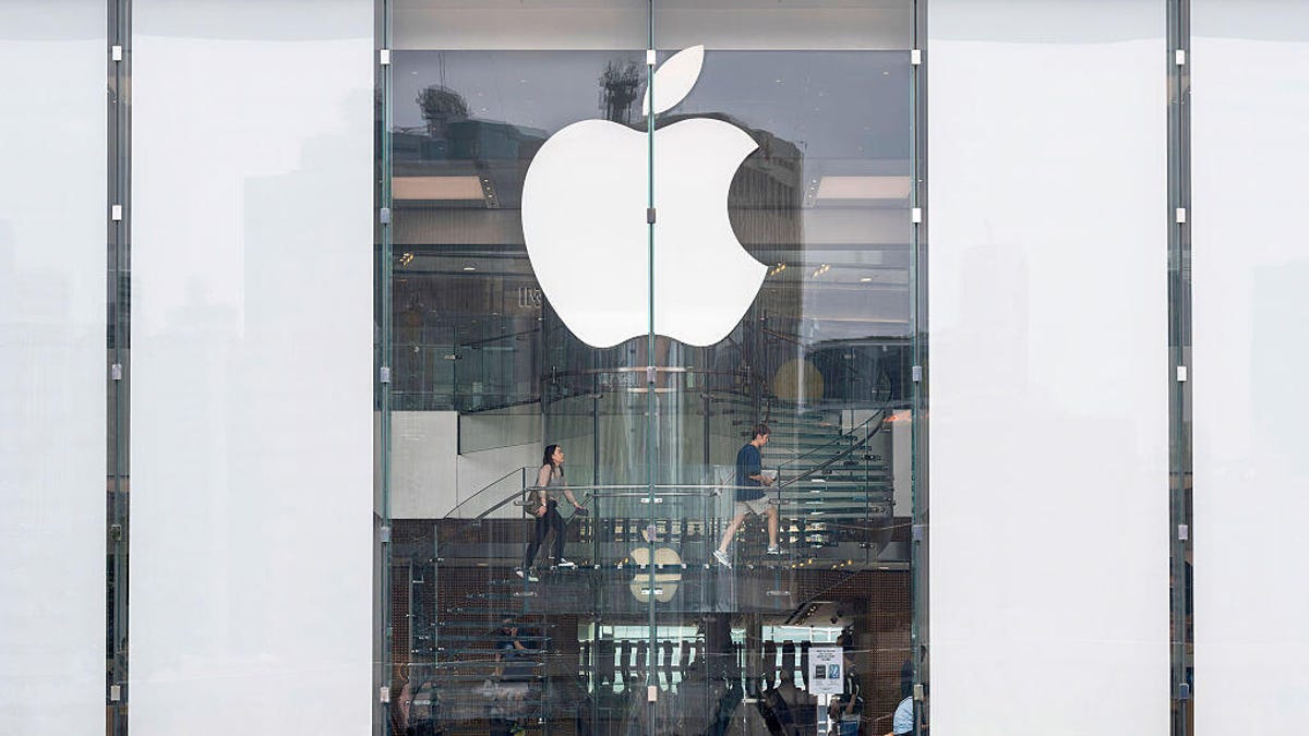 Shoppers walk up the stairs at an Apple Store in Hong Kong, with glass windows and the white Apple logo in the foreground with a crease in the middle.