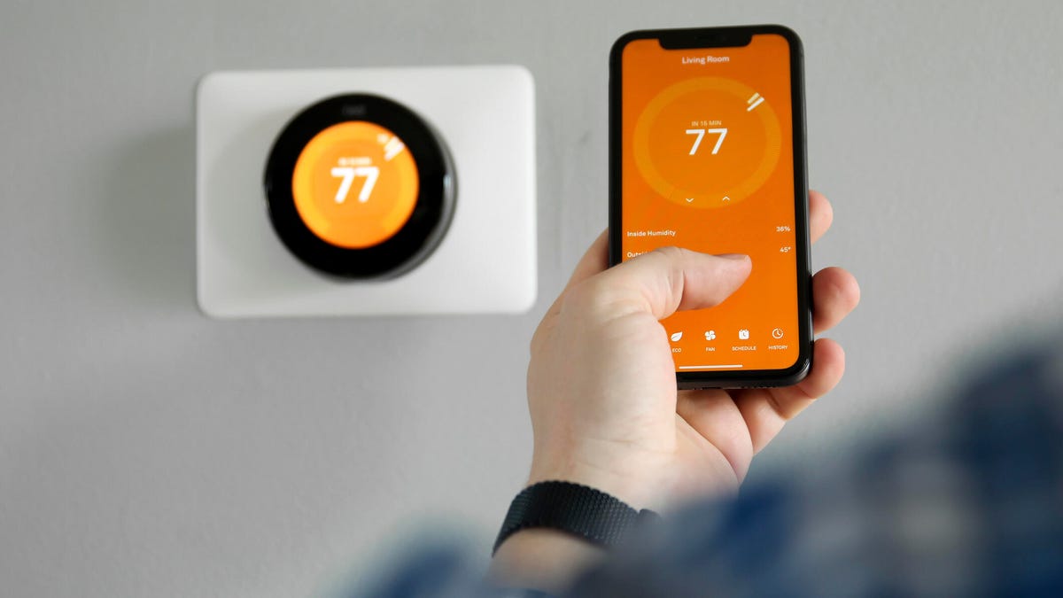 A person controls a thermostat from a phone, with an orange background on both the thermostat and the phone.