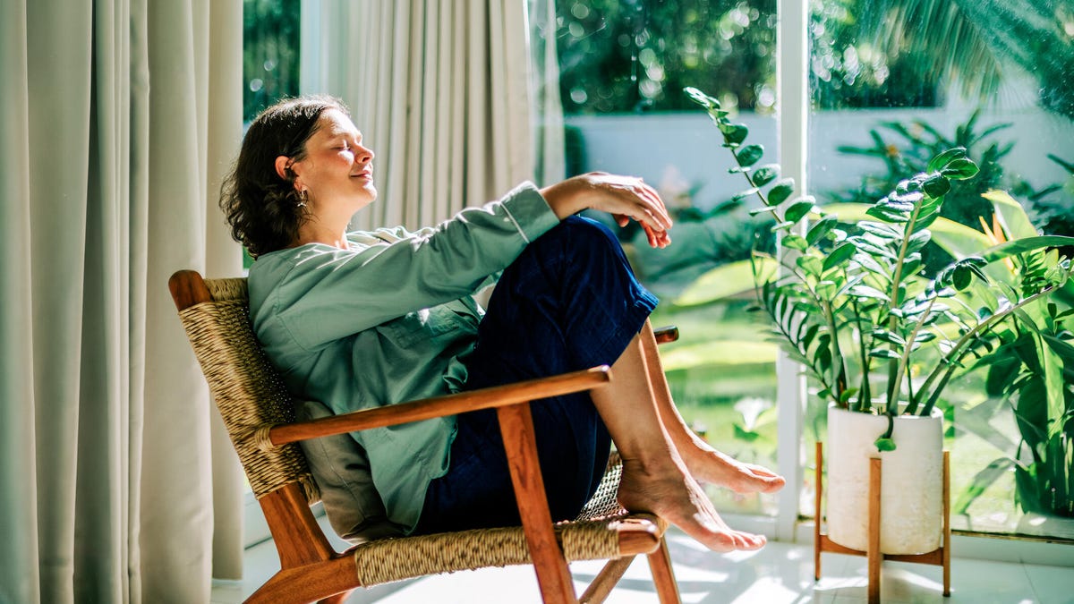 A woman sits in a chair in a sunroom and smiles.