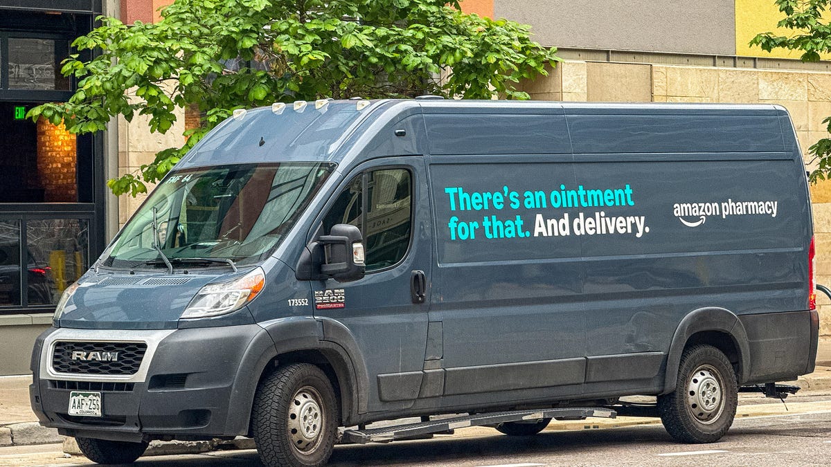 A dark blue Amazon Pharmacy van, parked on the side of the road in front of a short green tree. Text on the van reads, "There's an ointment for that. And delivery."