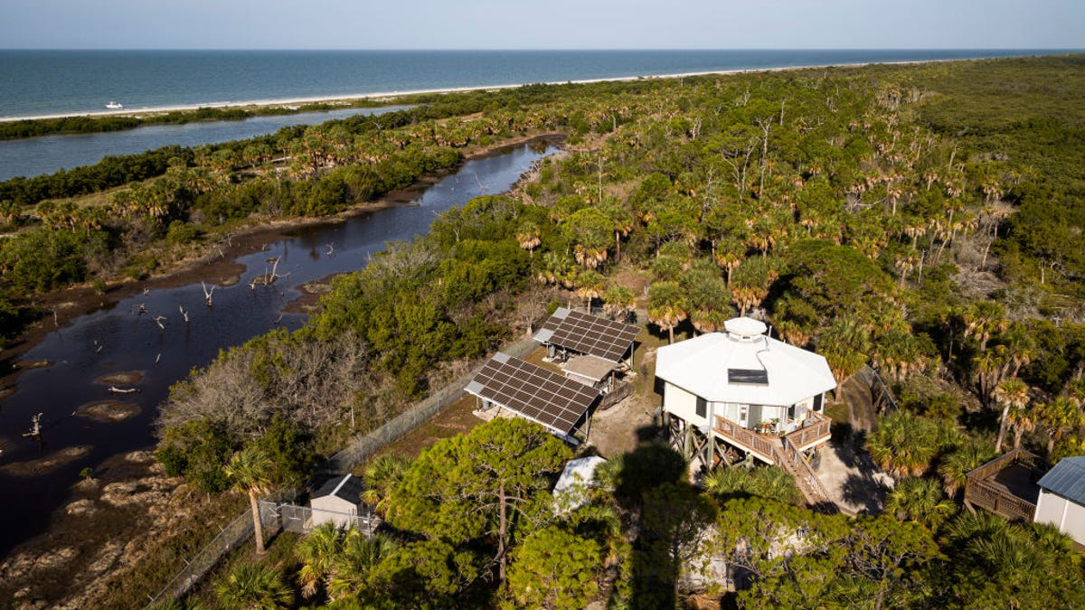 A house on a wooded island with two large arrays of solar panels next to it. The ocean and a beach are in the background.