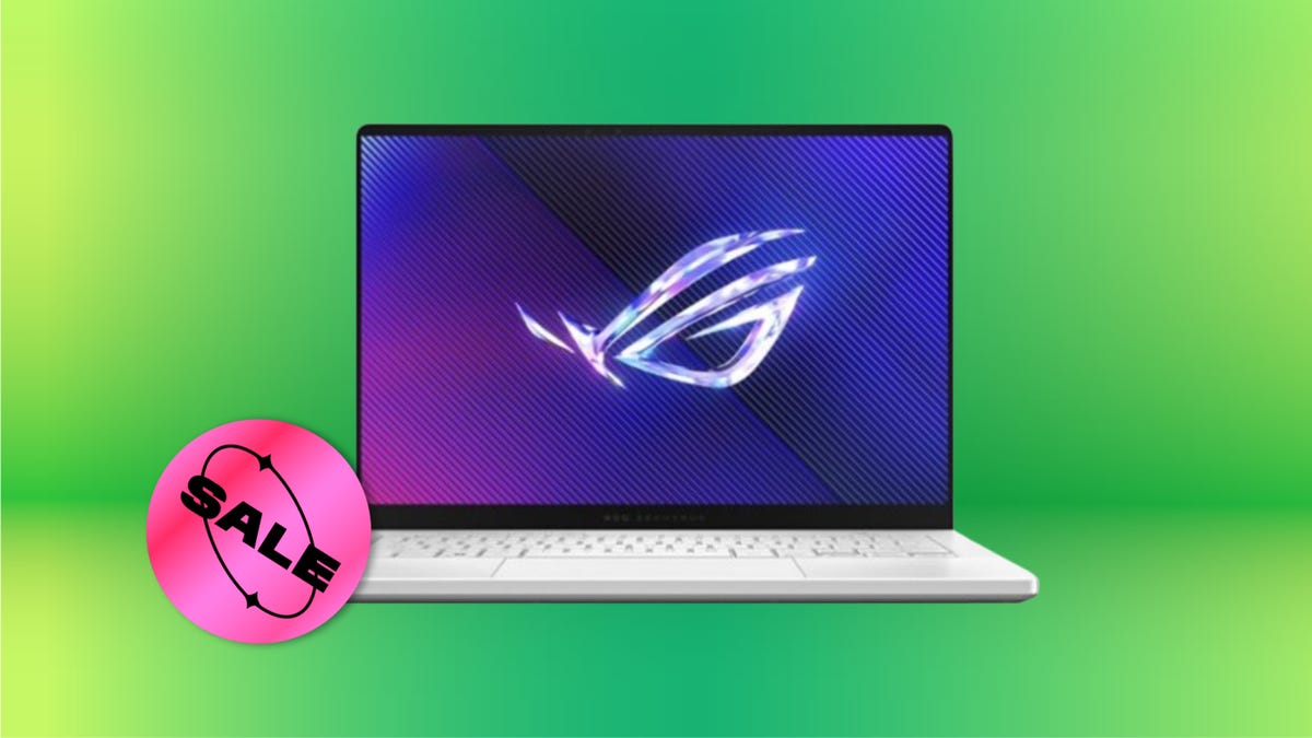 Silver colored laptop with purple wallpaper against green background. Circular pink "sale" sticker is at the lower left corner of the laptop.