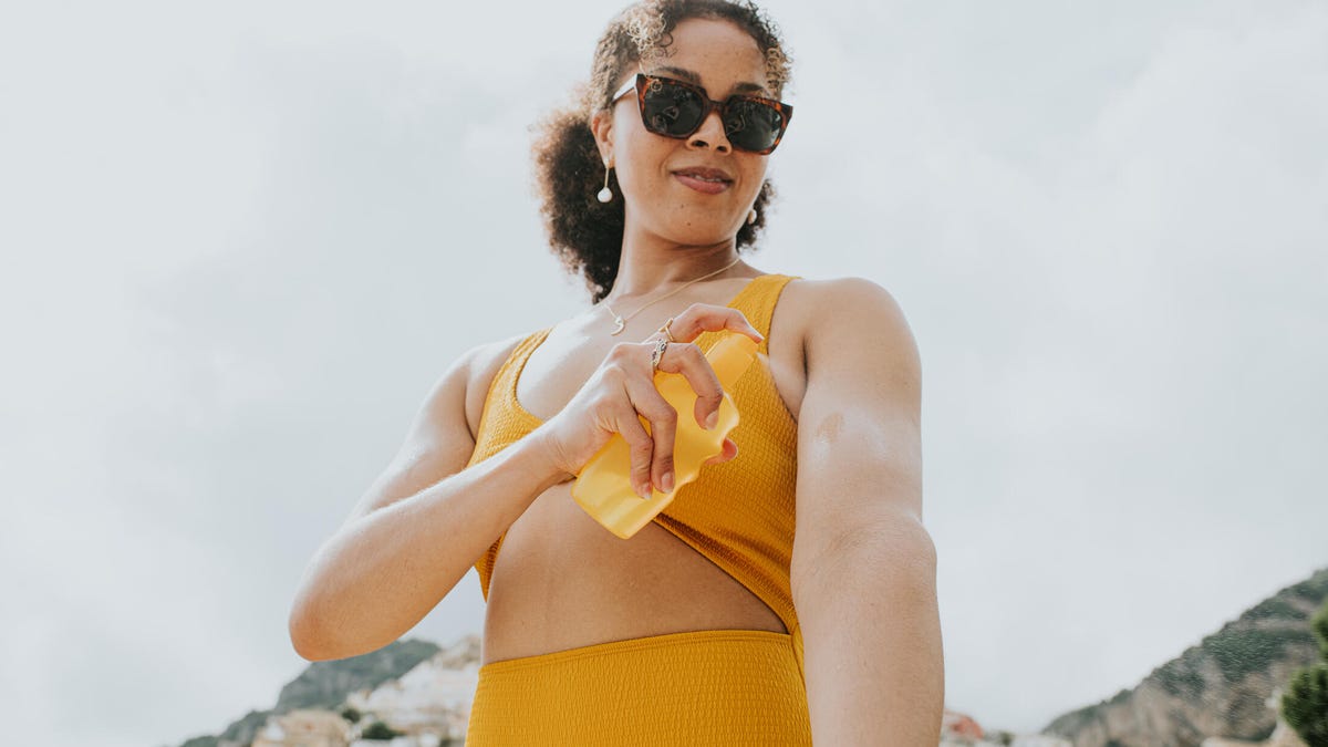 A person with curly brown hair in a ponytail, a yellow cut-out swimsuit, and sunglasses applying spray sunscreen on their arm.