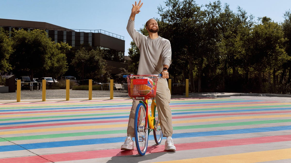 Steph Curry waving on a rainbow bike in a street paved with rainbow lines.