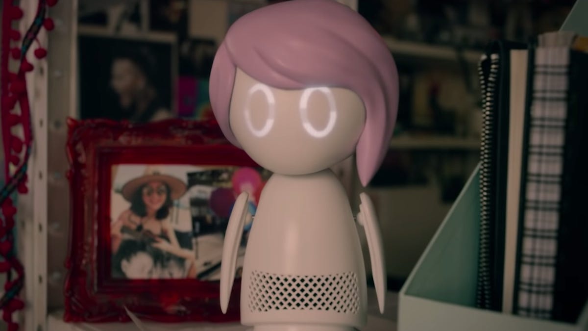 A small humanoid robot with glowing, ring-shaped eyes and molded pink "hair" stares at the camera. It looks a bit like a smart speaker with a head and small, penguin-like arms.