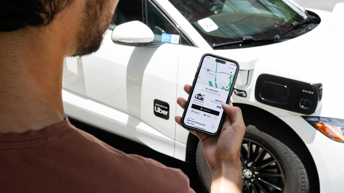 A man uses the Uber app on his phone as a Waymo Jaguar I-Pace pulls up in the background