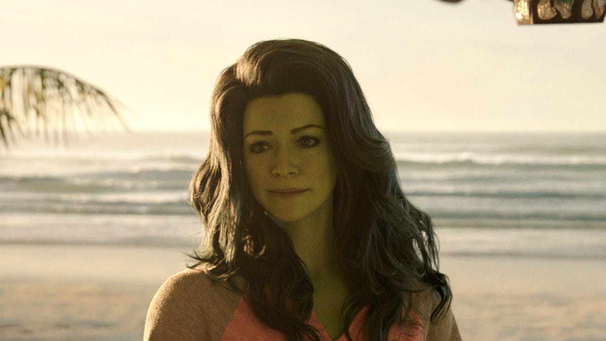 A green-skinned woman with long hair stands in front of the ocean.