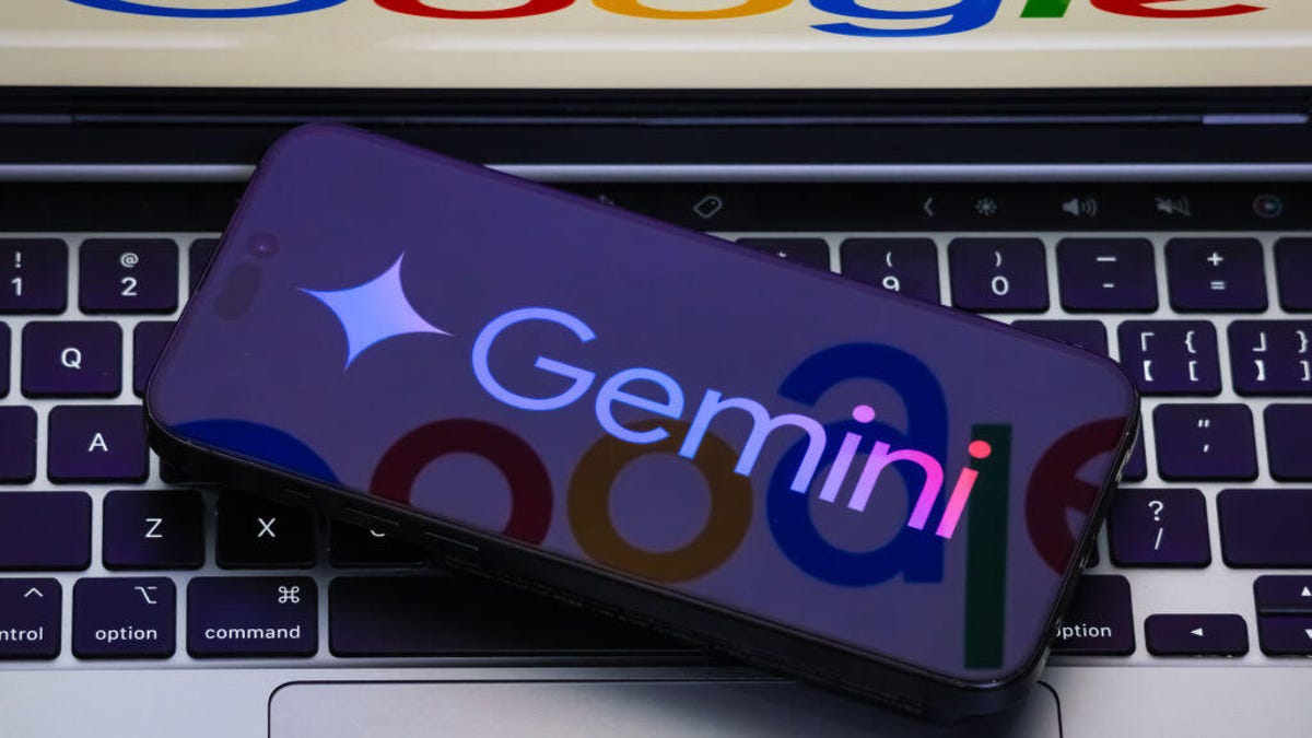 A phone displaying the Gemini logo lays on a keyboard of a laptop that has the Google logo on it.