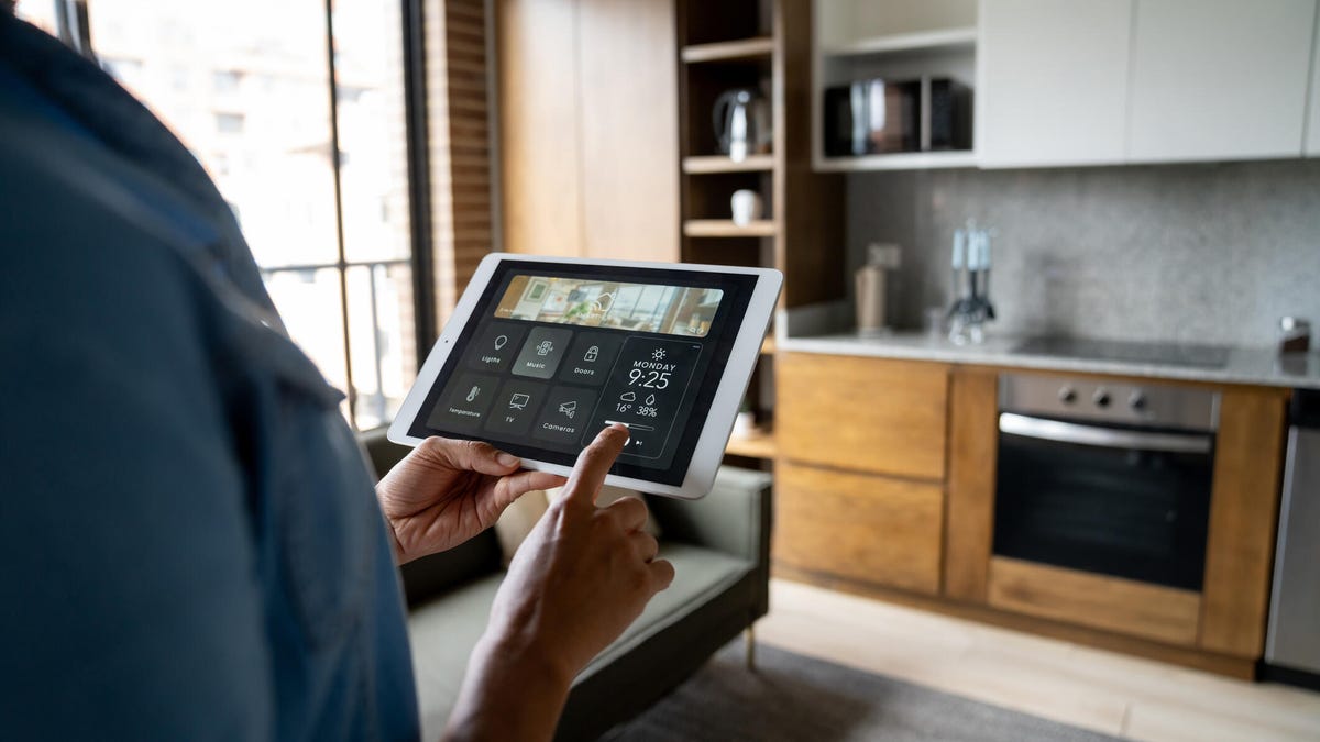 A person holds a tablet with smart home tabs in a sunlit kitchen.