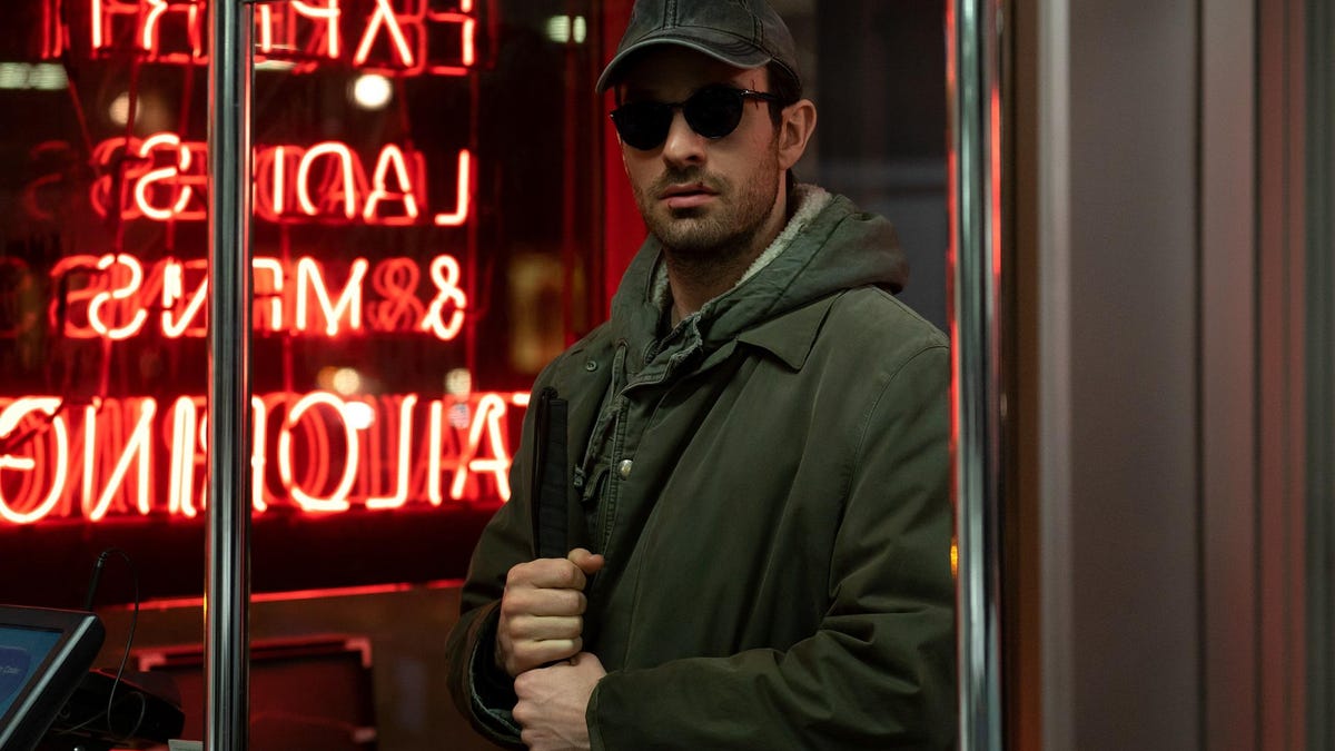 The character of Daredevil stand in front of a neon sign holding his walking cane.
