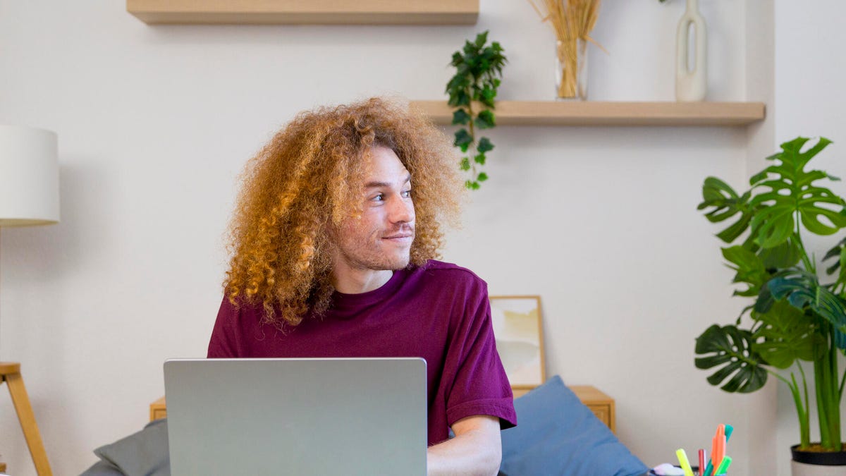 Thoughtful young man with curly hair working from home office looking away from laptop screen.