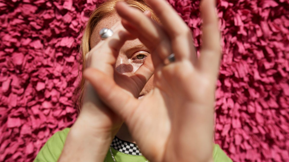A woman standing against a pink wall using her hands a telescope