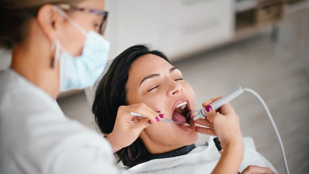 A dental hygienist with pulled-back blonde hair, glasses and a blue face mask removing calculus from a patient's teeth with a scaling tool. The patient has dark brown hair.