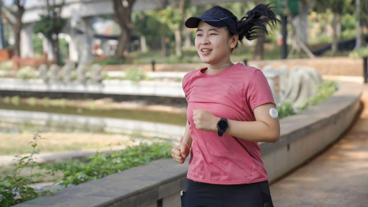 A person jogging in a pink tee, black visor, continuous glucose monitor on their arm and a black watch in a park setting.