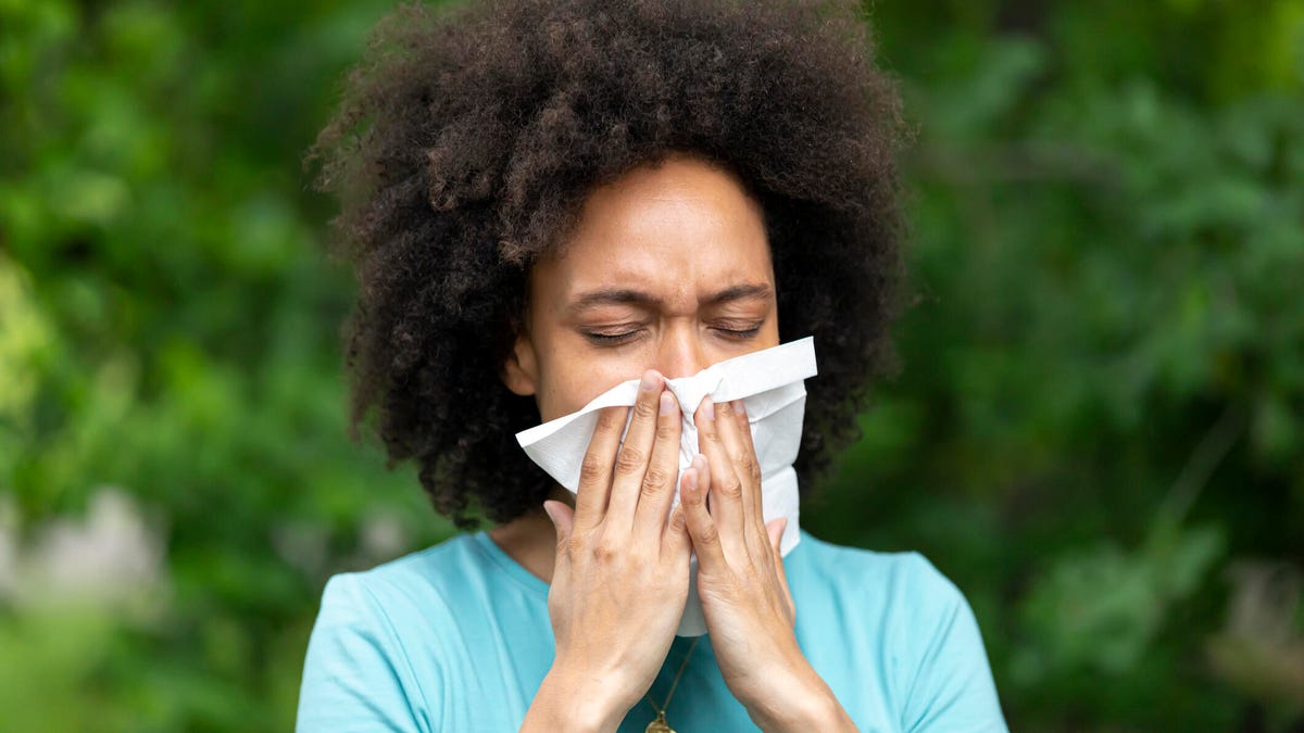 A person with curly brown hair and a bright blue tee blowing their nose with green trees in the background.