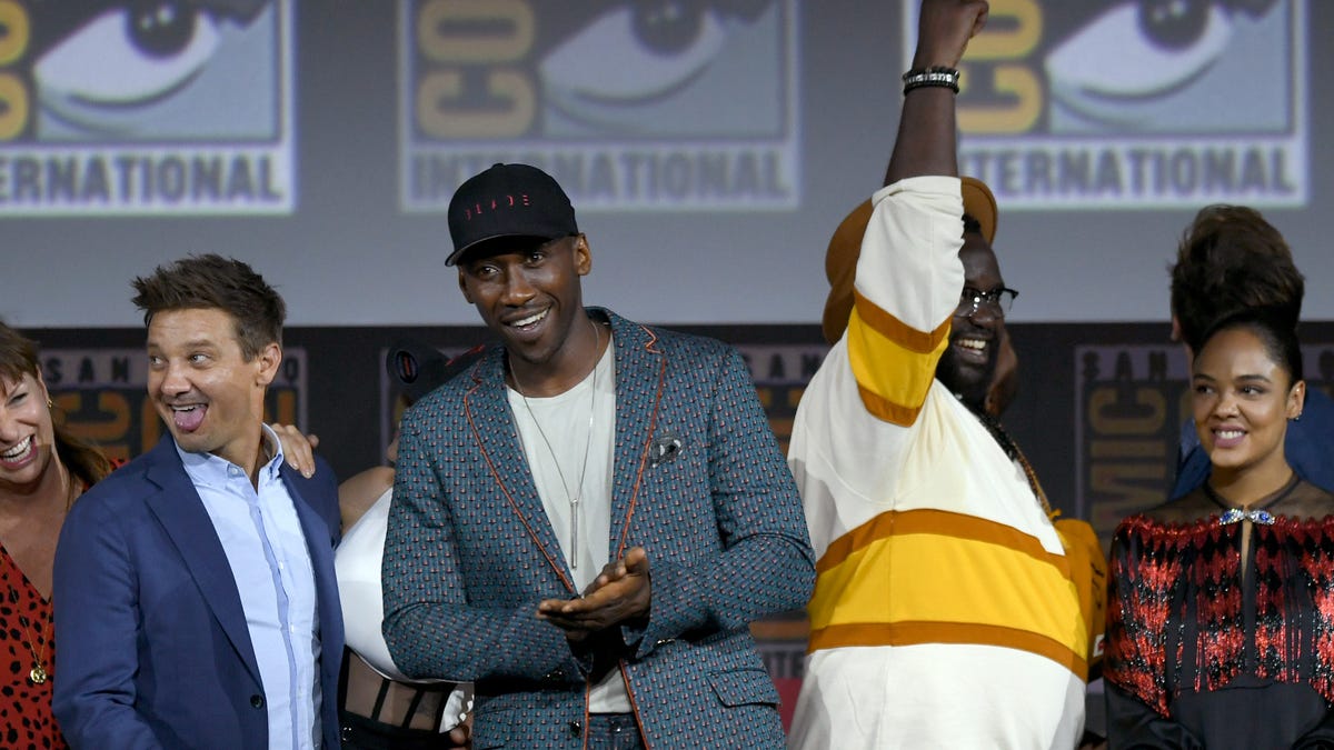 Actor Mahershala Ali in a grey suit and baseball cap smiles on stage at Comic Con, surrounded by other Marvel movie stars.
