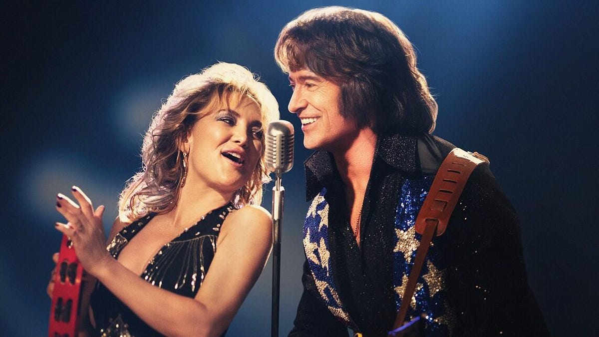 A blonde woman sings and plays a tamborine next to a man that looks like Neil Diamond.