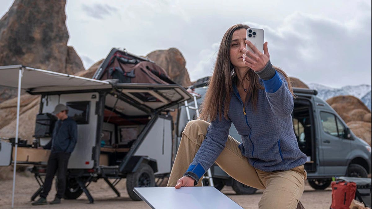 woman holding a phone and a rectangular satellite dish, kneeling in front of a camper van