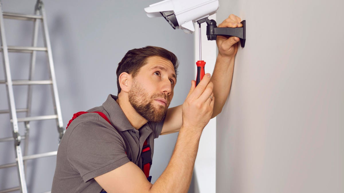 A person looks up at a security camera on the wall while adjusting it with a screwdriver.