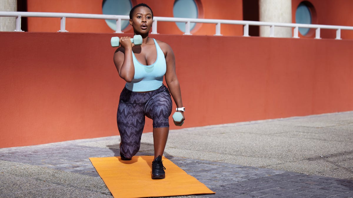 A person wearing a light blue exercise top and purple patterned leggings lifting light blue weights on a bright yellow yoga mat outside on concrete.