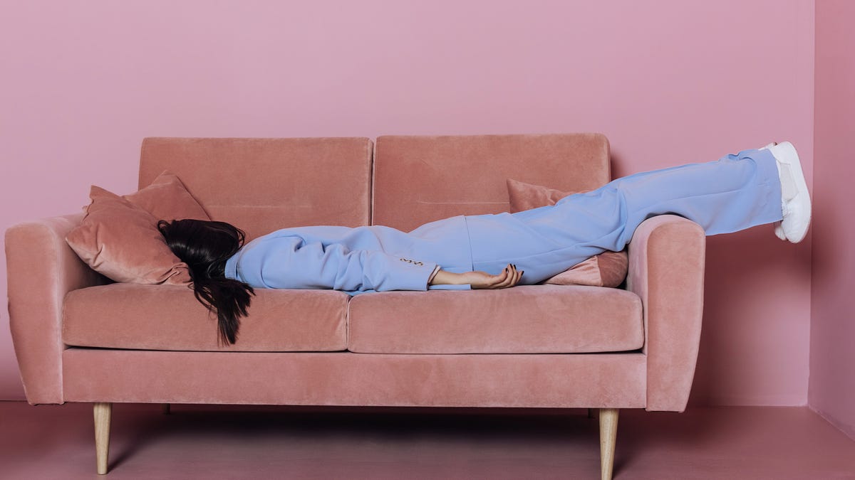 A person in a light blue suit lying face-down on a pink velvet sofa in a pink room.