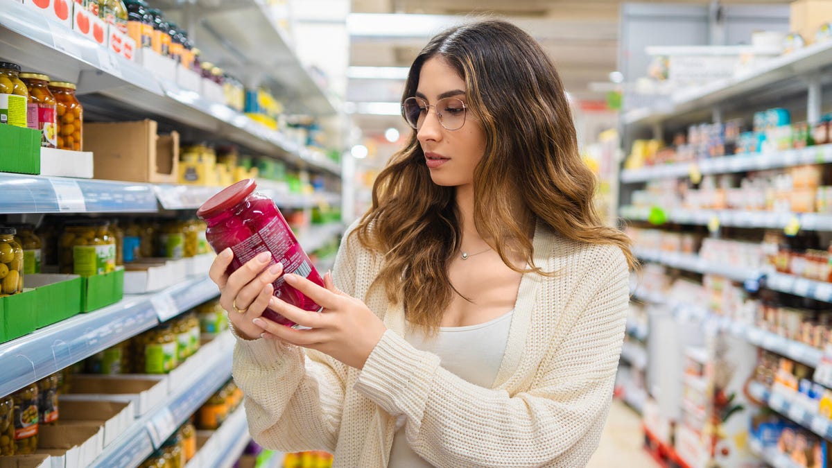 A photo of a woman reading the label on a jar of food in the grocery store.