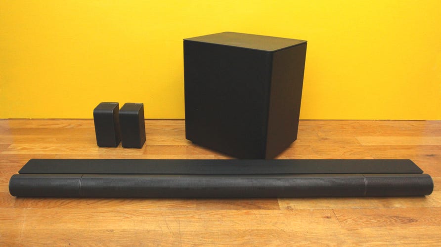 The Vizio Elevate soundbar, subwoofer, and dual rear speakers on a wooden floor with a yellow background.