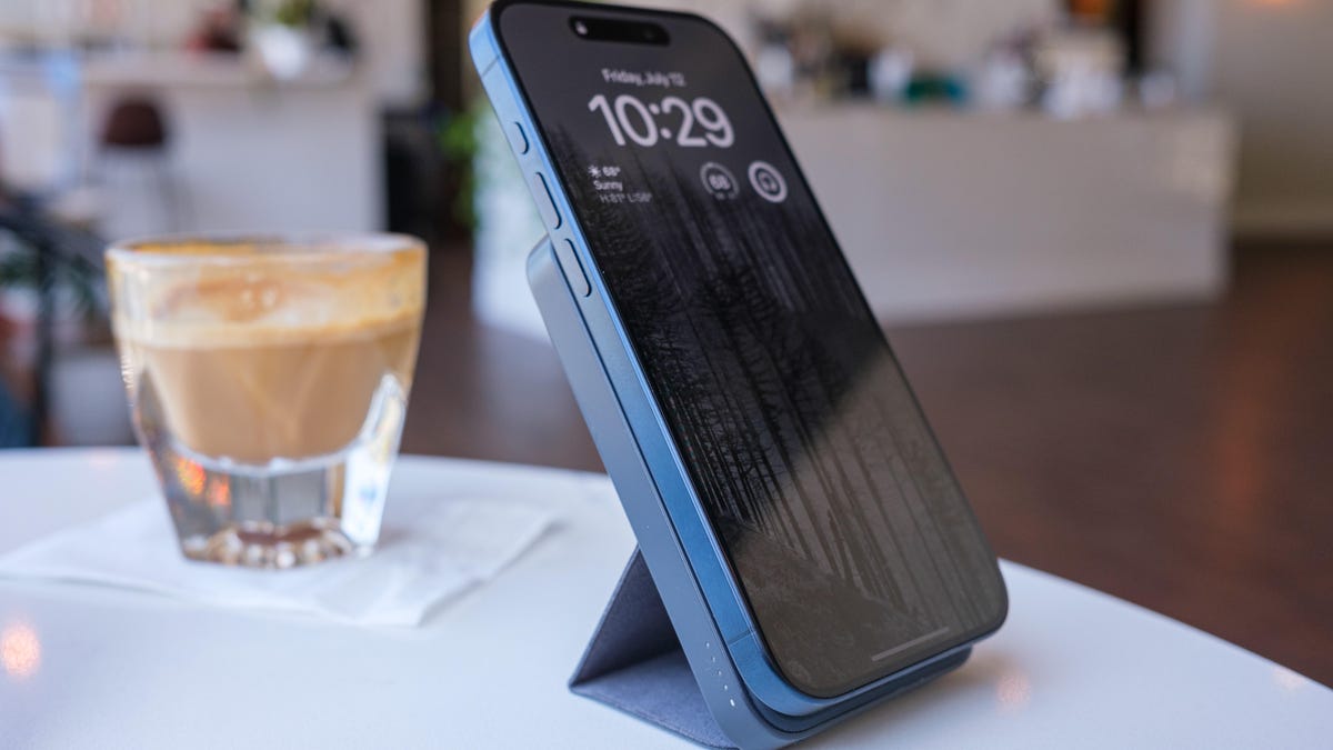 iPhone connected to a slim portable charger on a stand. They're both on a table in a brightly lit cafe along with a cortado in a glass.