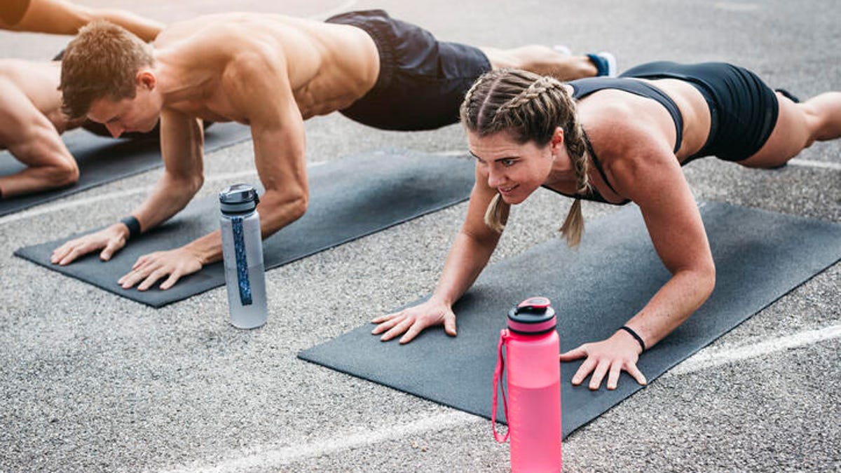 A young man and woman planking outside over their yoga mats with their water bottles in the foreground.