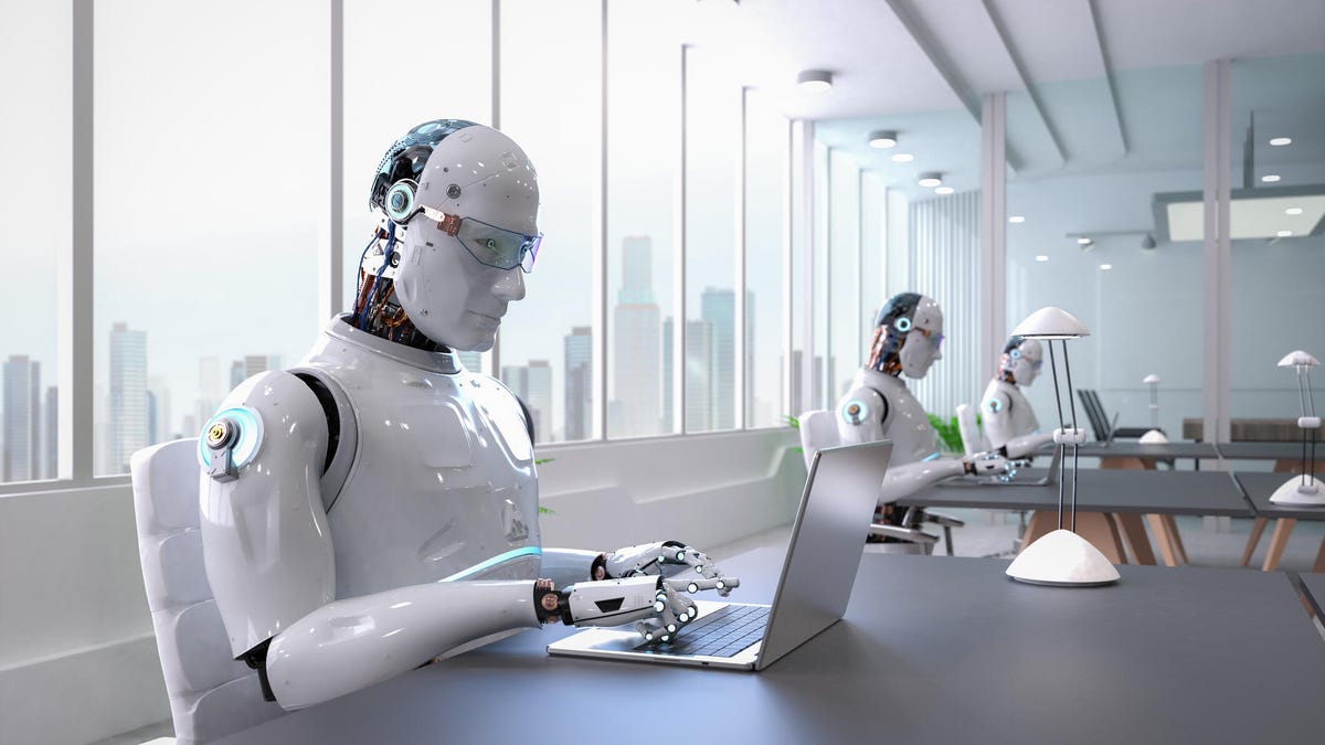 Humanoid robots working on laptops at tables in an office setting.