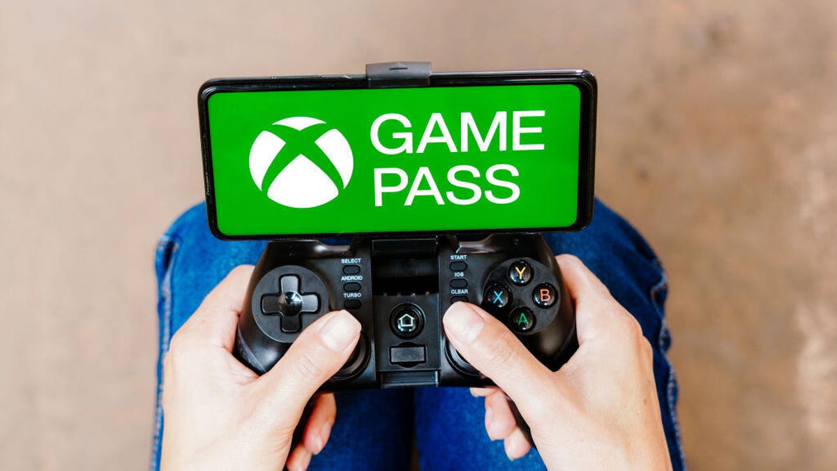 A person holding a game controller connected to a smartphone. The Xbox Game Pass logo is on the smartphone's screen