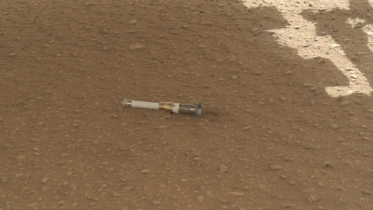 Mars sample tube resembles the handle of a Star Wars lightsaber. It's on the Martian ground, which is dusty and pebbly, and in the shadow of the rover.