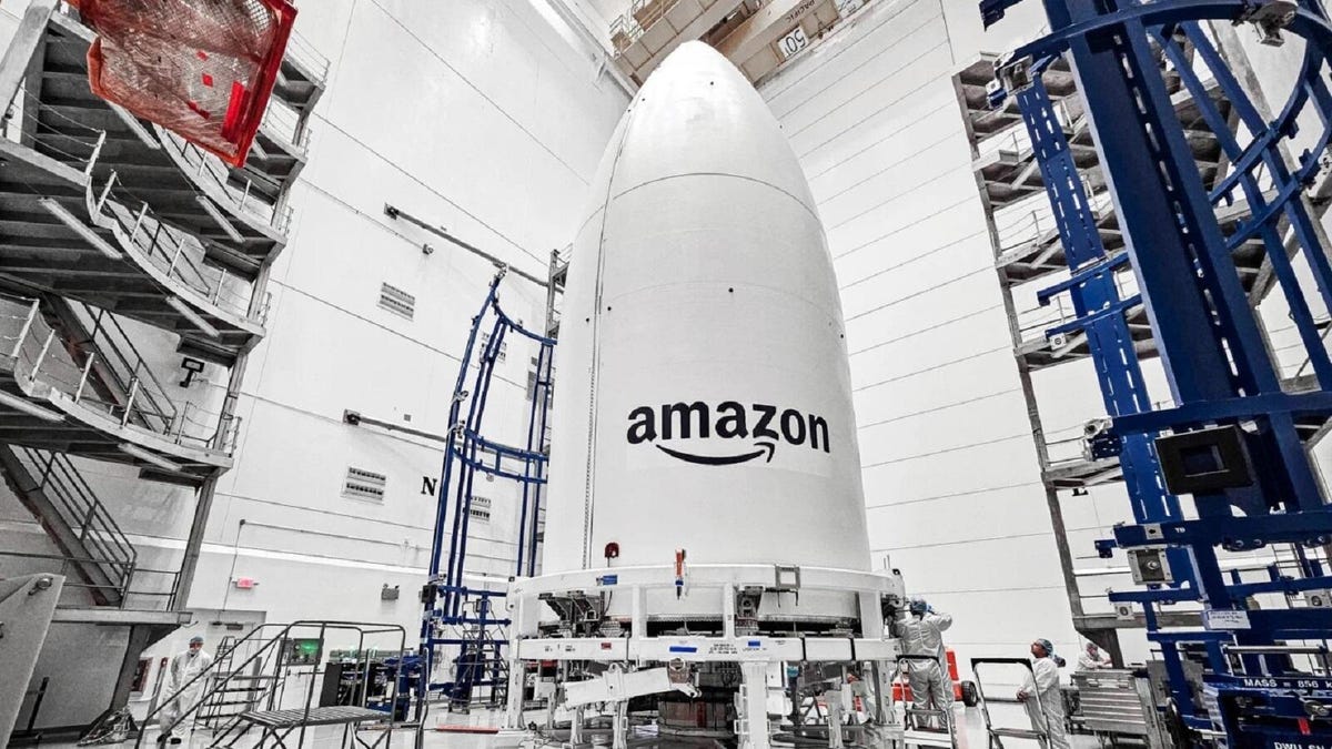 A large rocket payload with a big Amazon logo complete with smile arrow, planned to place atop an Atlas V rocket, sits in a pre-launch hanger.
