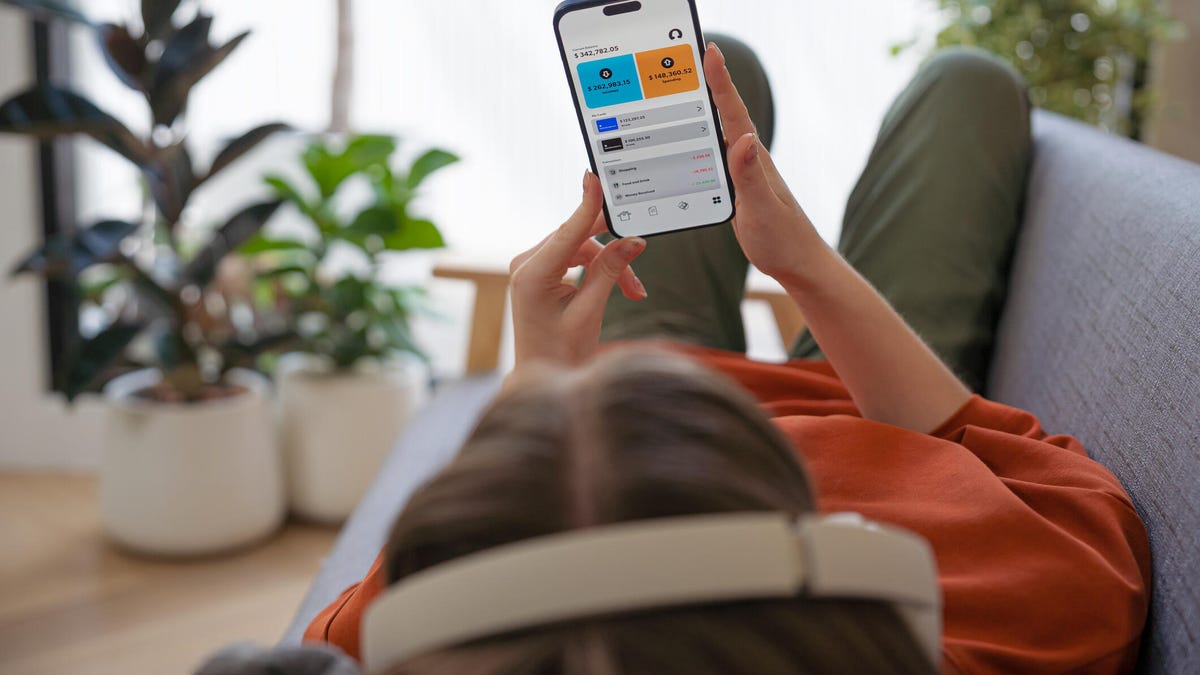 A woman in headphones lies on a sofa looking at a phone app at home.