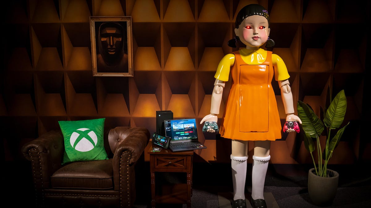A six-foot-tall replica of a doll wearing an orange dress over a yellow shirt. A pillow with the Xbox logo is on a chair