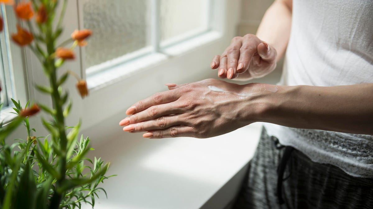 A person applying sunscreen on their hand indoors in front of a window next to a plant with orange flowers.