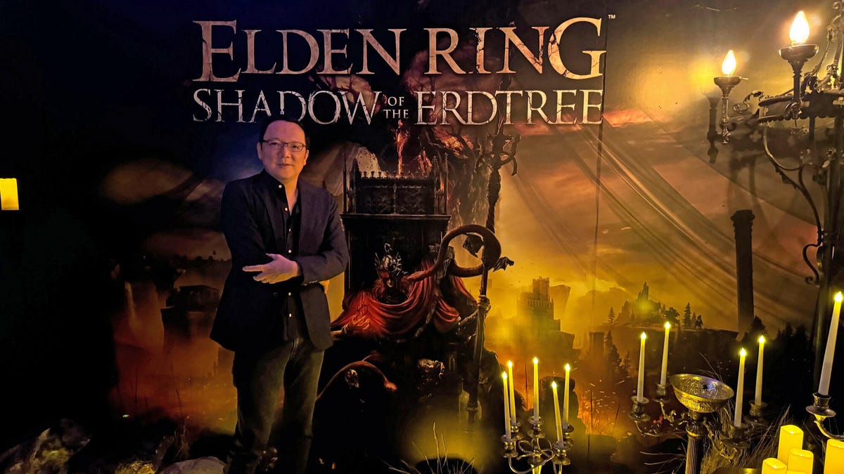 A man (and myth and legend), Hidetaka Miyazaki, stands in front of a wall-spanning poster for the Elden Ring DLC, Shadow of the Erdtree.