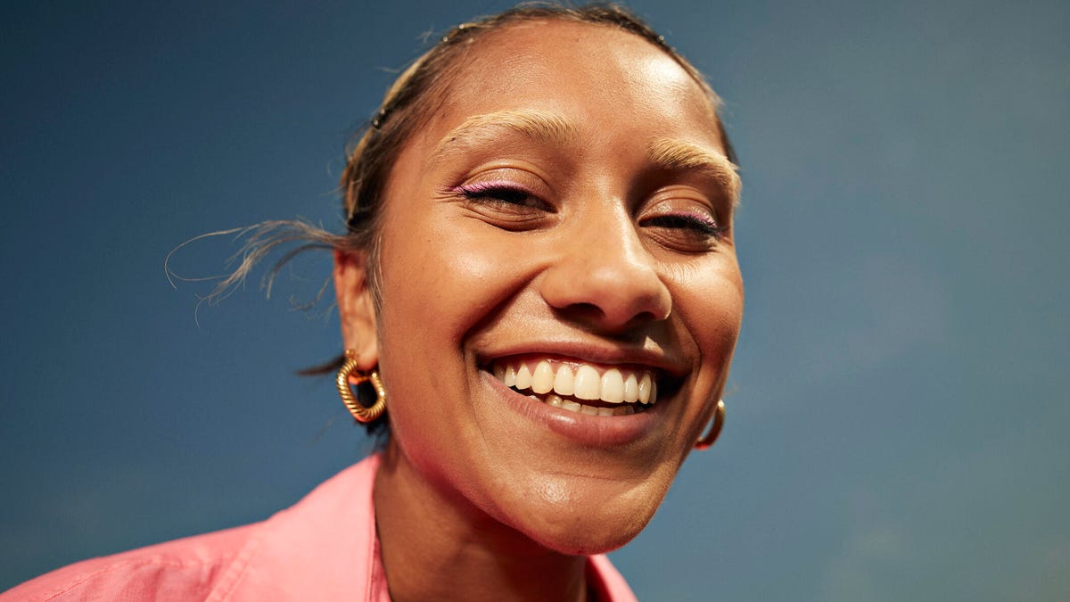 A person smiling in a pink shirt with gold hoop earrings and pulled-back hair.